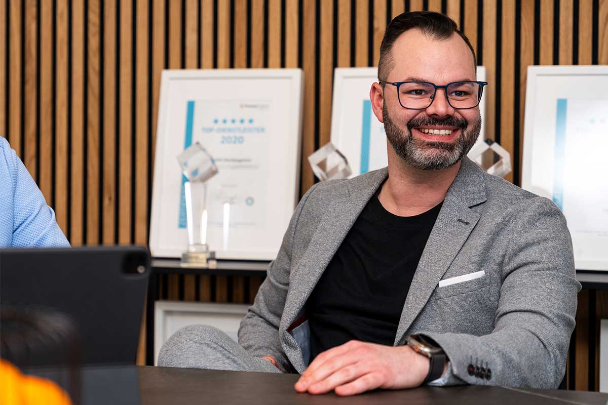 Porträt eines lächelnden Mannes in grauem Anzug mit schwarzem Shirt, der an einem Meeting teilnimmt. Im Hintergrund sind Auszeichnungen und Zertifikate der MAPO Werbeagentur Potsdam zu sehen.